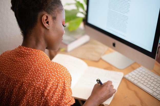 woman writing in her notebook sitting in front of a computer monitor
